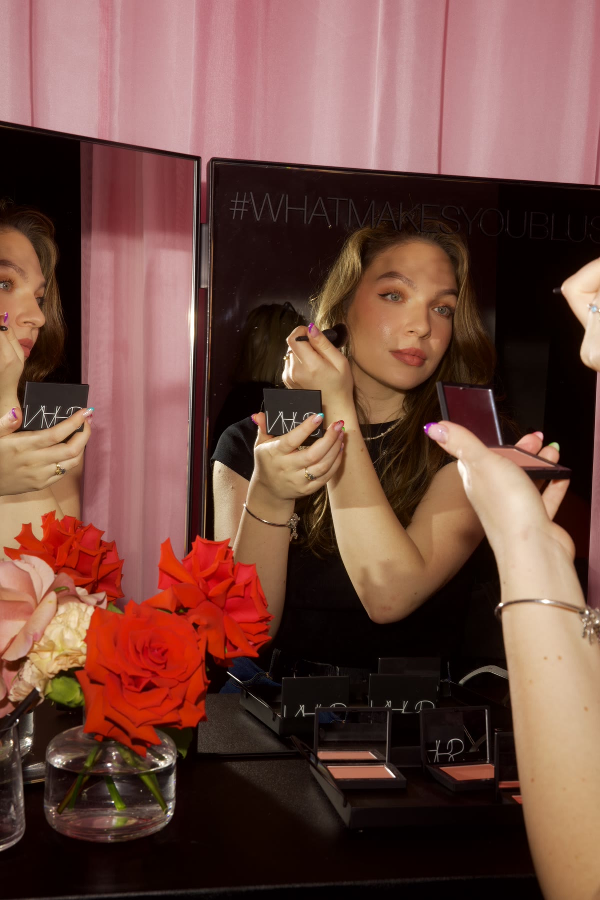Kasia applying blush at a mirrored vanity surrounded by roses and NARS products.